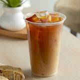 20 oz plastic cup filled with iced coffee on white table, cookie on napkin beside it, potted plant on wooden tray in background