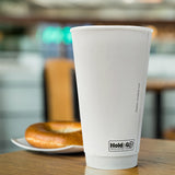 20 oz double-walled paper cup with "Hold & Go" text, on wooden table, next to tea plate with bagel, cafe in background