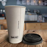 20 oz double-walled paper cup with "Hold & Go" text, next to black dome lid, on wooden table, cafe bar with menu and dark grey wall in background
