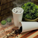 Two 20 oz cups on patterned table, one upright with coffee and foam art, one on side with spilled beans, next to potted plant and cactus candles, fenced wall