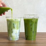 Two plastic cups with iced matcha on wooden table, left cup with liquid being poured into it, right cup with sip lid, white background
