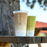 Two 24 oz plastic cups, left with iced coffee with milk, right with iced matcha with milk, on wooden bench, street and tree in background