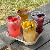 Four 24 oz plastic cups, filled with iced fruit drinks with fruit chunks, on cardboard coffee carrier, on wooden table, grass in background