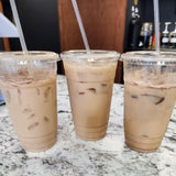 Three 24 oz plastic cups with flat lids and clear straws, filled with iced milk coffee, on white granite table, cafe in background