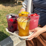 Four 24 oz plastic cups, filled with iced fruit drinks with fruit and berry chunks, in cardboard cup carrier held by person in striped shirt and brown pants, grass and curb outside