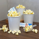 Three 32 oz paper food containers, filled with popcorn, with a scoop and movie tickets next to them on a wooden table