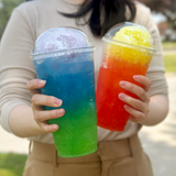 Two 32 oz plastic cups with dome lids, filled with colorful slushy drinks, held by person in beige shirt and brown pants, outside