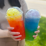 Two 32 oz plastic cups with dome lids, filled with colorful slushy drinks, held at an angle by person in beige shirt and brown pants, grass in background
