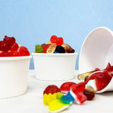 Three 5 oz paper food containers filled with variety of gummy candy, on white table, blue wall in the background