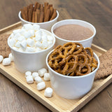 Four 8 oz paper food containers, filled with cinnamon, pretzels and marshmallows, on a wooden tray on a table
