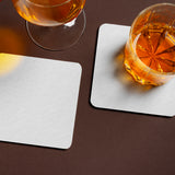 Two 4" Blank, White Medium Weight Square Coaster, with a whiskey glass resting a on top and a wine glass to the side, on a brown background