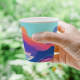 10 oz. Multicolor Recyclable Paper Cup with a wave design a pattern of various colours and shapes, posed with a hand, outdoors.