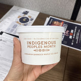 12 oz paper food container, with "Indigenous People Month" text print, held by a hand, with a wooden table and posters in the background