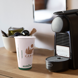 16 oz compostable paper cup with green band and "ecotainer" text, brown "Cabin" logo with trees and moon, on wooden table beside coffee machine with coffee pods