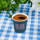 4 oz. Full Wrap Recyclable Paper Cup displaying "I like coffee", beside flowers, on a blue, plaid table cloth.