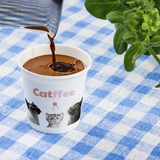 4 oz. Multicolor Recyclable Paper Cup with "Catffee" logo, with espresso being poured, on a blue plaid table cloth, next to a plant.