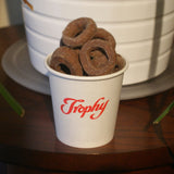 Custom printed 4 oz paper cup with red "Trophy" logo, filled with chocolate snacks, on dark wooden table, plant pot in background