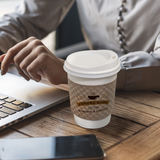White paper coffee cup with a kraft brown sleeve featuring "Coffee Shop" logo, positioned near a person using a laptop.