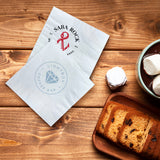 Two 3-PLY White Beverage Napkin of different logos lying slightly apart, beside fruit cake and a bowl of melted chocolate topped with marshmallows, on a wooden table