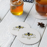 Three white round coasters, with "Oats & Honey Cafe" logo and a print of bee, on white table, next to jar of lemon and glass of tea.