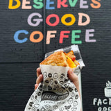 White paper sandwich bag featuring the "Factory Coffee" logo, holding a sandwich, displayed in a hand against a black brick wall.