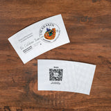 Two white coffee cup sleeves showing the front and back designs featuring "The Kraken" logo on one side and a QR code on the other, on a wooden table