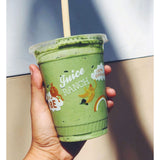 24 oz plastic cup with flat lid and straw, filled with green smoothie, "Juice Ranch" logo with illustrations, held by hand, beige and white wall background