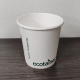 4 oz compostable paper cup with green "ecotainer" text, on grey wooden table, white wall background