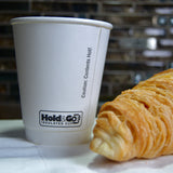 8 oz double-walled paper cup with "Hold & Go" text, on white napkin next to croissant, tiled wall in background